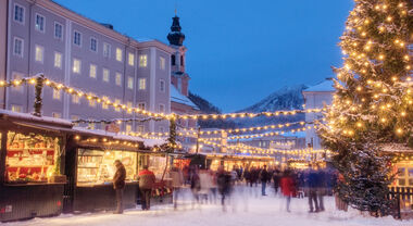 Verschneiter Weihnachtsmarkt bei Abenddämmerung mit Besuchern und beleuchtetem Weihnachtsbaum